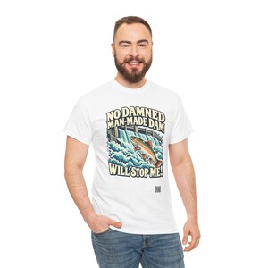 Puede incluir: Una camiseta blanca de manga corta con un gr&aacute;fico de un pez saltando sobre una presa y el texto "NO DAMNED MAN-MADE DAM WILL STOP ME!" impreso en letras oscuras y negritas sobre y debajo de la ilustraci&oacute;n del agua y el pez.