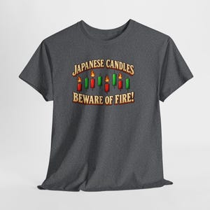 May include: A dark heather grey short-sleeve t-shirt featuring a graphic of red and green Japanese candlestick chart bars with flames. The text reads "JAPANESE CANDLES" above the bars and "BEWARE OF FIRE!" below in a bold, light-coloured font.