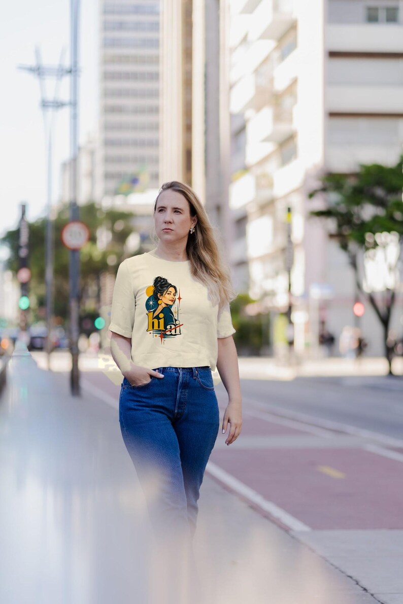 May include: A cream-colored short-sleeve t-shirt featuring a graphic illustration of a person with dark hair in a bun, accompanied by the text "hi" in bold yellow letters, set against a background of blue circles and orange starburst accents.