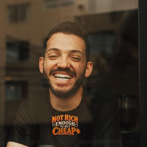May include: A black short-sleeved t-shirt featuring the orange and white text "NOT RICH ENOUGH TO BUY CHEAP" printed on the chest. The shirt is worn by a person with a beard who is laughing while sitting behind a white coffee cup with a black lid.
