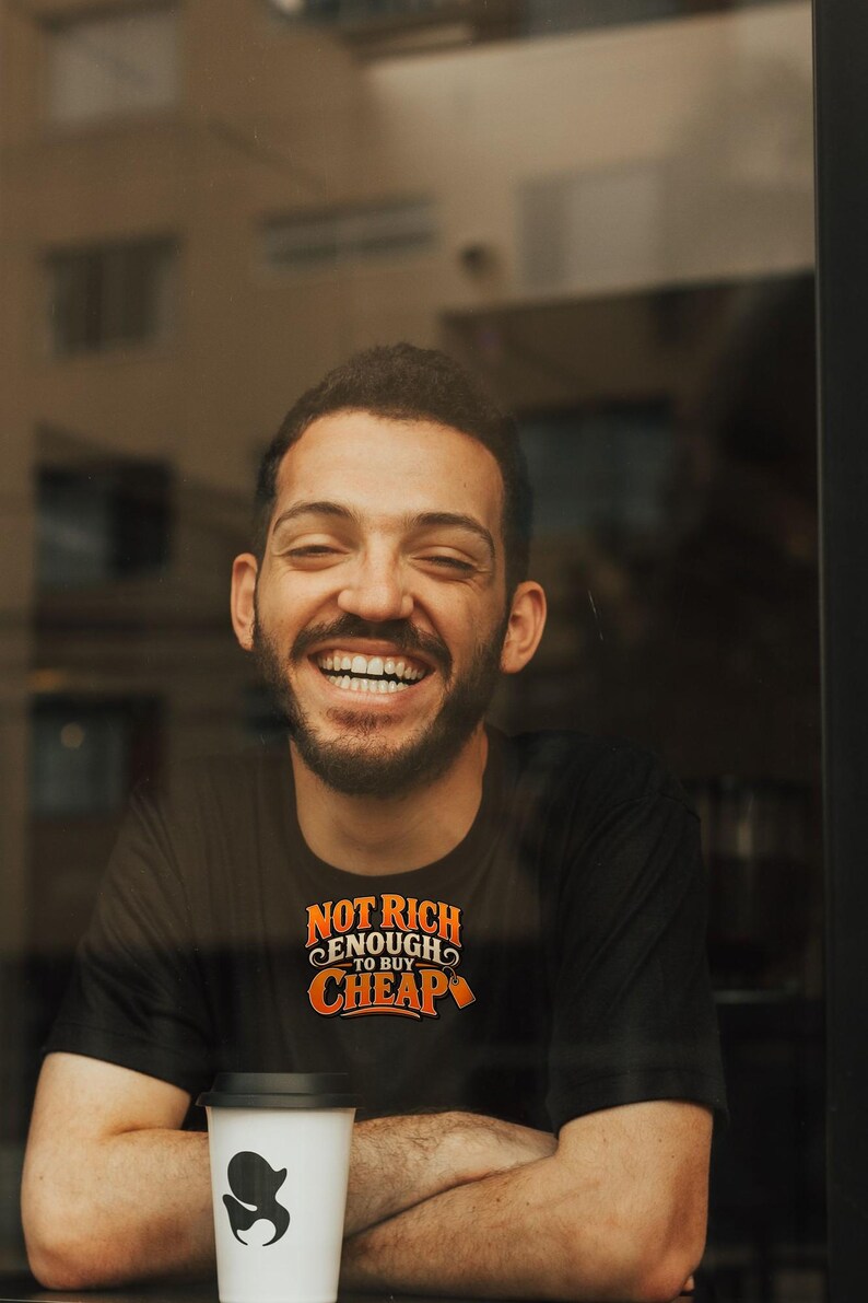 May include: A black short-sleeved t-shirt featuring the orange and white text "NOT RICH ENOUGH TO BUY CHEAP" printed on the chest. The shirt is worn by a person with a beard who is laughing while sitting behind a white coffee cup with a black lid.