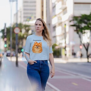 May include: A light blue short-sleeved t-shirt featuring a graphic illustration of a tan Pomeranian dog on the chest, with the word Pomeranian printed in black text above the dog's head, paired with dark blue denim jeans.