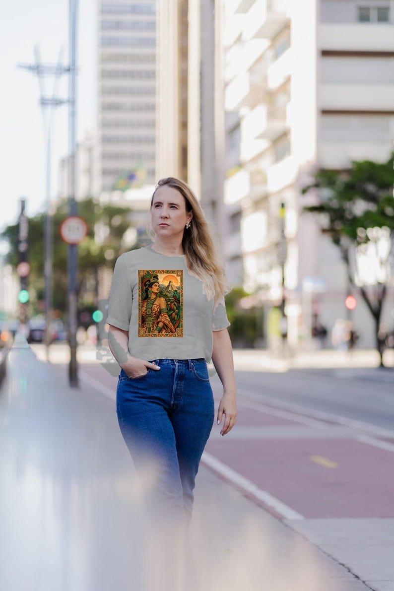 May include: A light gray short-sleeved t-shirt featuring a rectangular graphic print of a person wearing ornate traditional attire and a feathered headdress, set against a backdrop of a sun and lush green foliage in warm orange and green tones.