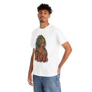 May include: A white t-shirt featuring a graphic print of an Aztec warrior wearing a large green feathered headdress, a gold chest plate, and red and green patterned clothing. The warrior is depicted in profile with detailed ornaments.
