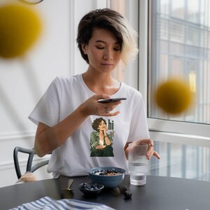 Op de afbeelding: Een wit T-shirt met korte mouwen en een grafische print van een persoon met krullend haar in een groen overhemd en een donkere broek, met de kin op de hand rustend. De tekst "hi ai" staat in een donkergroen lettertype naast de afbeelding.