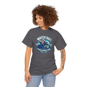 May include: A dark gray t-shirt featuring a graphic of a pilot relaxing in a cockpit with a robot co-pilot. The text reads WATCH OUT! I'M ON AUTOPILOT NOW. The design includes blue and white circular accents with wings and a small airplane icon.
