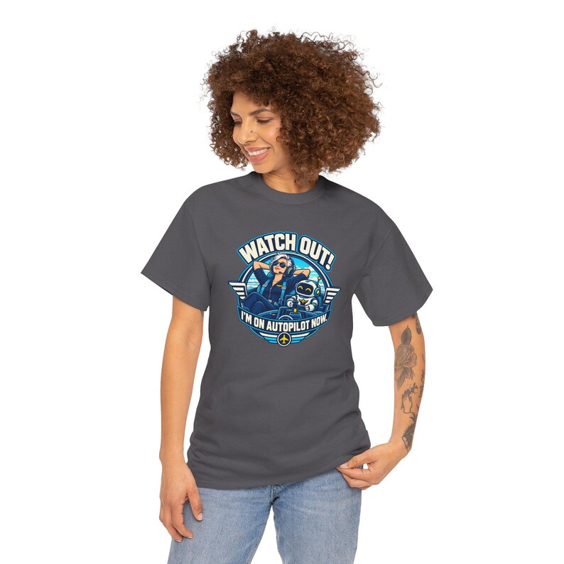 May include: A dark gray t-shirt featuring a graphic of a pilot relaxing in a cockpit with a robot co-pilot. The text reads WATCH OUT! I'M ON AUTOPILOT NOW. The design includes blue and white circular accents with wings and a small airplane icon.
