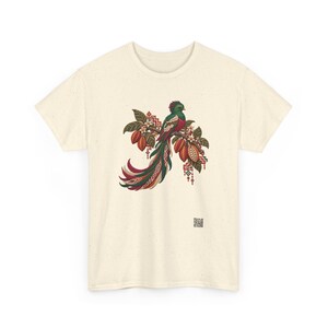 May include: A cream-colored short-sleeve t-shirt featuring a central graphic of a quetzal bird perched on a branch with cacao pods and leaves. The bird has vibrant green, red, and white plumage with long tail feathers and geometric patterns.