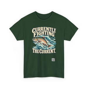 May include: A dark green t-shirt featuring a graphic of a fish jumping through blue and white waves. The text above the fish reads CURRENTLY FIGHTING and the text below reads THE CURRENT. A small white square QR code is printed near the bottom.