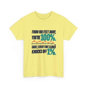 May include: A pale yellow short-sleeve t-shirt featuring dark blue and teal text that reads: FROM 100 FEET AWAY, YOU'RE 100%. SADLY, EVERY FOOT CLOSER KNOCKS OFF 1%. The design includes a horizontal line with small orange footprints.