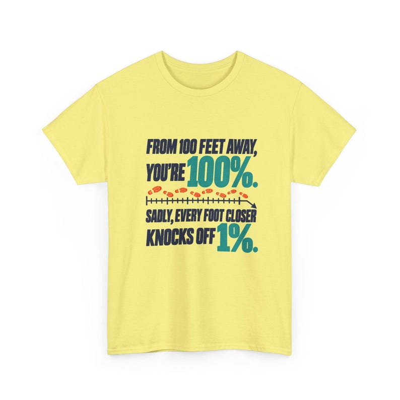 May include: A pale yellow short-sleeve t-shirt featuring dark blue and teal text that reads: FROM 100 FEET AWAY, YOU'RE 100%. SADLY, EVERY FOOT CLOSER KNOCKS OFF 1%. The design includes a horizontal line with small orange footprints.