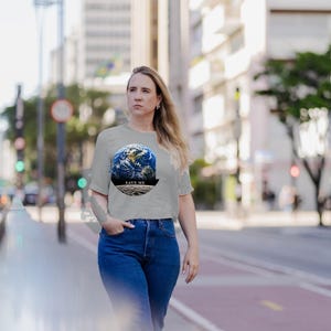 May include: A light gray short-sleeved t-shirt featuring a circular graphic of the Earth above a lunar surface. The text "SAVE ME" is printed in black capital letters within a dark banner positioned below the Earth and above the moon.
