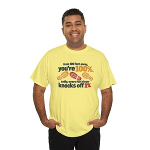 May include: A light yellow short-sleeve t-shirt featuring the text 'from 100 feet away, you're 100%. sadly, every foot closer knocks off 1%.' with three shoe print graphics in orange and red colors centered on the chest area.