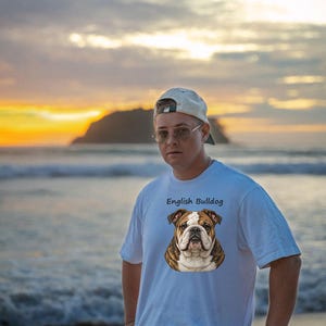 Puede incluir: Una camiseta blanca de manga corta con una ilustraci&oacute;n gr&aacute;fica de la cara de un bulldog ingl&eacute;s marr&oacute;n y blanco centrada en el pecho, con el texto English Bulldog impreso en letras negras justo encima de la ilustraci&oacute;n del perro.