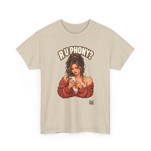 May include: A beige short-sleeve t-shirt featuring a graphic illustration of a person with long brown hair wearing a red cardigan, looking down at a smartphone. Above the figure, the text reads "R U PHONY?" in bold, arched letters.