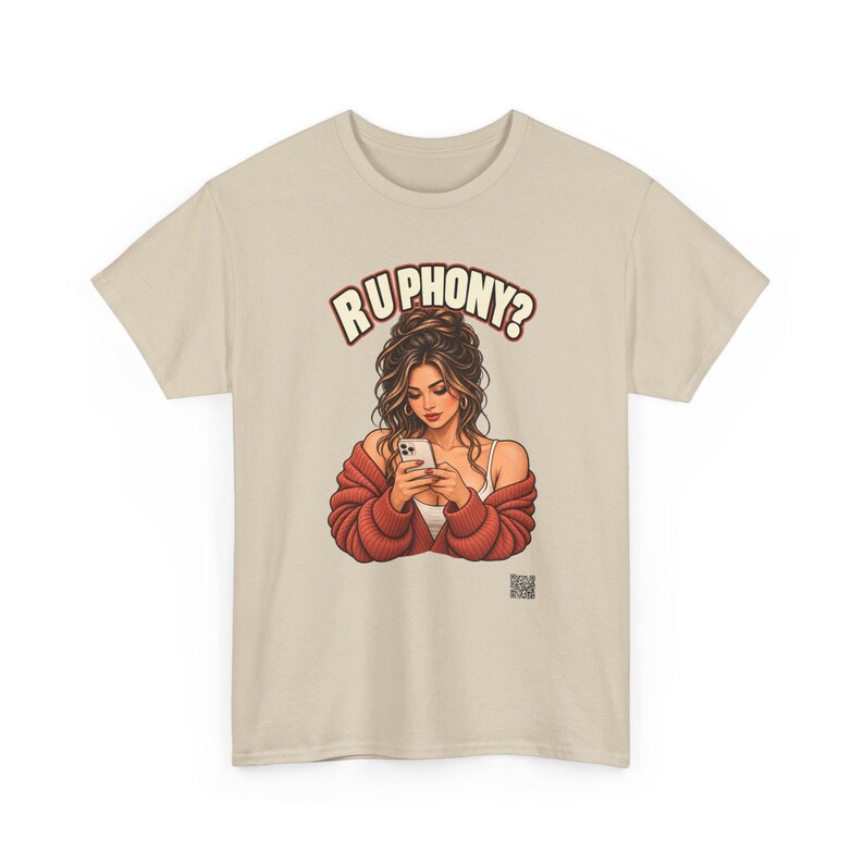 May include: A beige short-sleeve t-shirt featuring a graphic illustration of a person with long brown hair wearing a red cardigan, looking down at a smartphone. Above the figure, the text reads "R U PHONY?" in bold, arched letters.