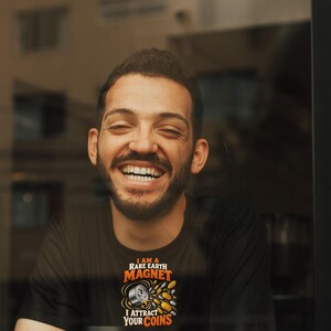 May include: A black t-shirt featuring a graphic of a smiling magnet with the text I AM A RARE EARTH MAGNET I ATTRACT YOUR COINS in orange and white lettering. The design includes small metallic coin illustrations surrounding the magnet.