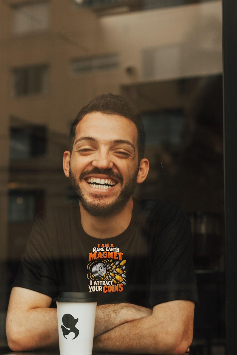May include: A black t-shirt featuring a graphic of a smiling magnet with the text I AM A RARE EARTH MAGNET I ATTRACT YOUR COINS in orange and white lettering. The design includes small metallic coin illustrations surrounding the magnet.