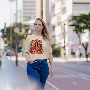 K&ouml;nnte beinhalten: Ein cremefarbenes Kurzarm-T-Shirt mit einem bunten Grafikdruck einer Person in traditioneller Kleidung und dem Text "COME TO BELIZE PLEASE", umgeben von Blumen- und Vogelmotiven, kombiniert mit einer blauen Jeans.
