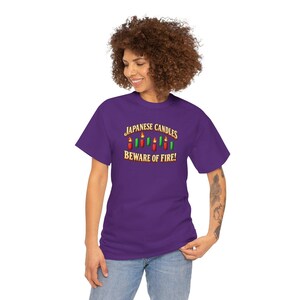 May include: A purple short-sleeve t-shirt featuring a graphic of red and green Japanese candle sticks with flames. The text reads JAPANESE CANDLES BEWARE OF FIRE! in a gold-coloured font. The shirt has a crew neckline and a standard fit.