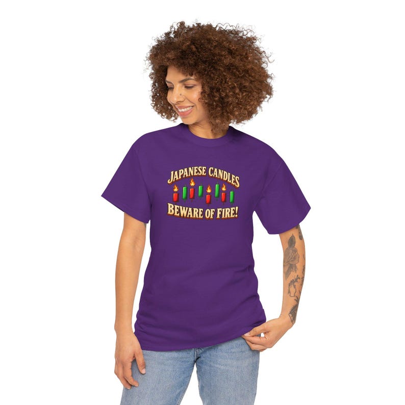 May include: A purple short-sleeve t-shirt featuring a graphic of red and green Japanese candle sticks with flames. The text reads JAPANESE CANDLES BEWARE OF FIRE! in a gold-coloured font. The shirt has a crew neckline and a standard fit.