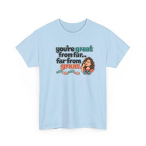 May include: A light blue t-shirt featuring a graphic with the text "you're great from far... far from great." The design includes a cartoon illustration of a person looking through binoculars and four small colorful footprints below the text.