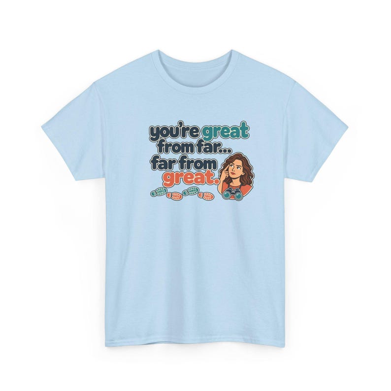 May include: A light blue t-shirt featuring a graphic with the text "you're great from far... far from great." The design includes a cartoon illustration of a person looking through binoculars and four small colorful footprints below the text.