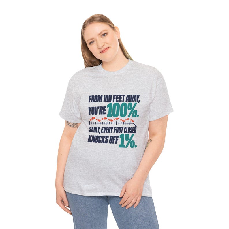 May include: A light gray short-sleeve t-shirt featuring dark blue and teal text that reads: FROM 100 FEET AWAY, YOU'RE 100%. SADLY, EVERY FOOT CLOSER KNOCKS OFF 1%. A graphic of a measuring line with small orange footprints is included.
