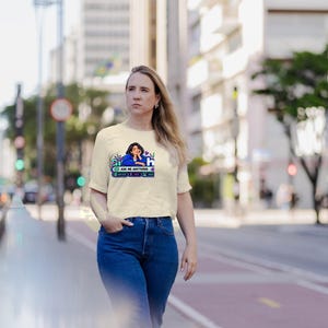 May include: A cream-colored short-sleeve t-shirt featuring a graphic print of a person in a blue shirt with the text "ASK ME ANYTHING" and various icons. The shirt is paired with blue denim jeans, creating a casual look with a clear, colorful design.