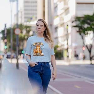 May include: A light blue short-sleeved t-shirt featuring a graphic of an orange cat sitting in a lotus position with the text "LESS DRAMA" above and "MORE KARMA" below the illustration, paired with dark blue denim jeans.