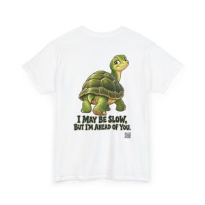 May include: A white t-shirt featuring a green turtle illustration and the text "I MAY BE SLOW, BUT I'M AHEAD OF YOU." The turtle is depicted with a dark green shell and light green skin, looking back over its shoulder. A small QR code is visible below the text.