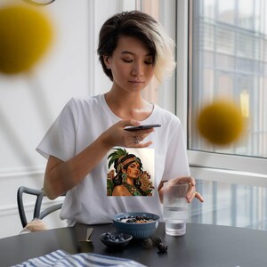 Puede incluir: Una camiseta blanca de manga corta con un estampado gr&aacute;fico de una persona de pelo oscuro, que lleva un tocado de plumas verdes y joyas ornamentadas, sobre un fondo de patrones geom&eacute;tricos y follaje tropical.
