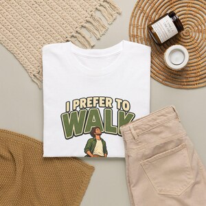 Op de afbeelding: Een wit t-shirt met de tekst I PREFER TO WALK in grote groene en beige letters boven een illustratie van een persoon met krullend haar in een groen overhemd en wit ondershirt, staand met gesloten ogen en het hoofd achterover gekanteld.
