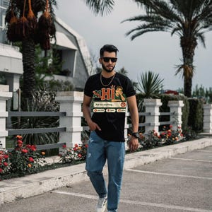 Puede incluir: Una camiseta negra de manga corta con un estampado gr&aacute;fico de una persona apoyada en una interfaz de chat con el texto "HI" y "Ask me anything..." combinada con vaqueros azul claro desgastados y zapatillas deportivas blancas.
