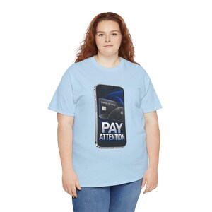 May include: A light blue t-shirt featuring a graphic of a smartphone displaying a black credit card labeled Bank of Mine. Below the card, the text PAY ATTENTION is printed in bold white letters. The design is centered on the chest of the shirt.
