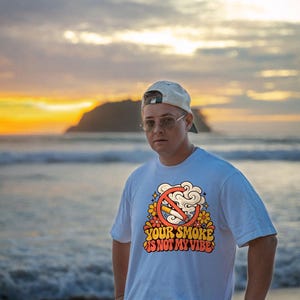 May include: A white short-sleeved t-shirt featuring a graphic print of a cigarette inside a red prohibition circle surrounded by orange and yellow flowers and smoke clouds, with the text "YOUR SMOKE IS NOT MY VIBE" in a retro font.