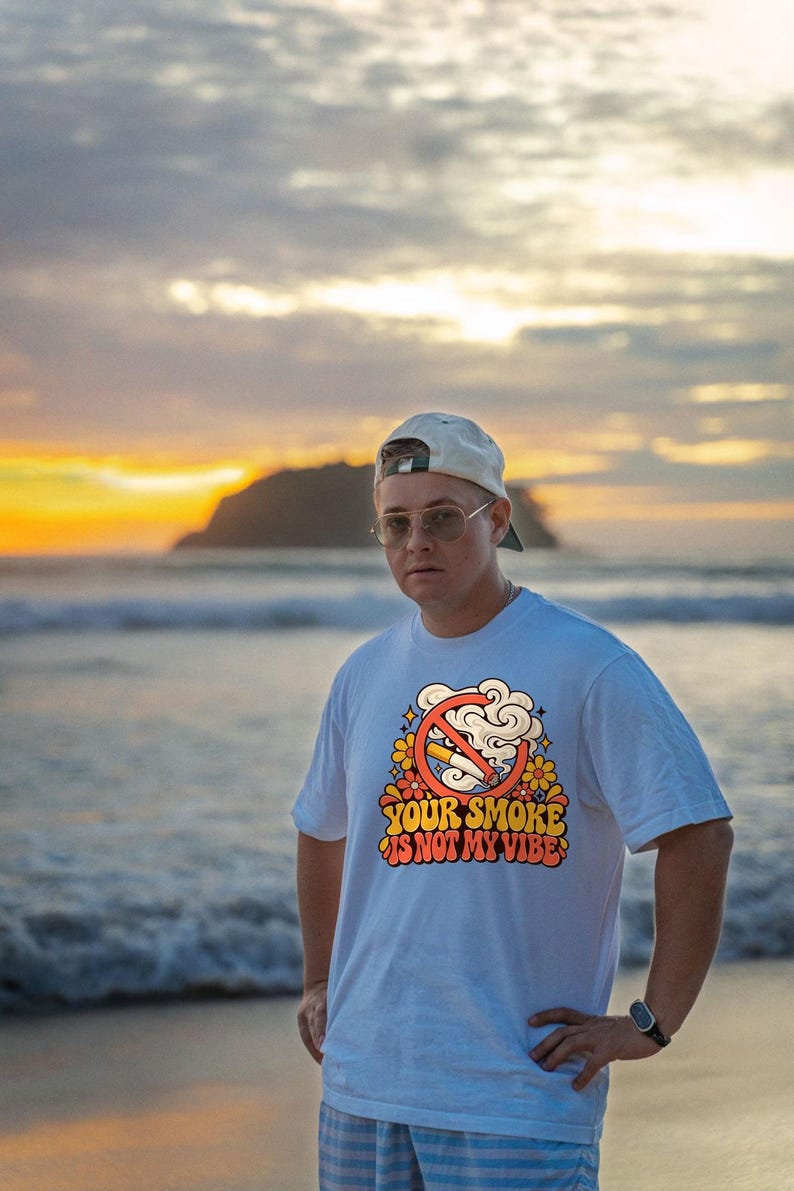 May include: A white short-sleeved t-shirt featuring a graphic print of a cigarette inside a red prohibition circle surrounded by orange and yellow flowers and smoke clouds, with the text "YOUR SMOKE IS NOT MY VIBE" in a retro font.
