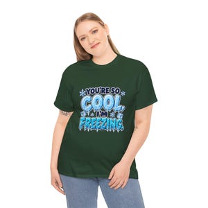 May include: A dark green short-sleeve t-shirt featuring a graphic design with the text "YOU'RE SO COOL, I'M FREEZING." The lettering is styled with an icy, blue, and white frozen texture, accented by small snowflakes and ice cubes.