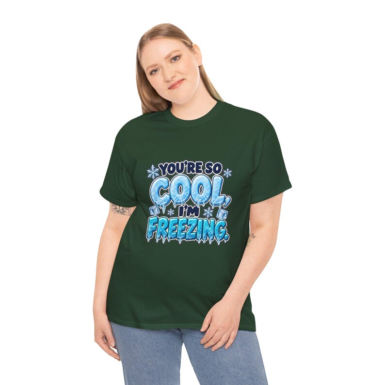 May include: A dark green short-sleeve t-shirt featuring a graphic design with the text "YOU'RE SO COOL, I'M FREEZING." The lettering is styled with an icy, blue, and white frozen texture, accented by small snowflakes and ice cubes.