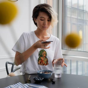 May include: A white short-sleeved t-shirt featuring a colorful graphic print of an Aztec warrior wearing a traditional feathered headdress and ornate jewelry. The shirt has a crew neckline and is made of a plain white fabric.