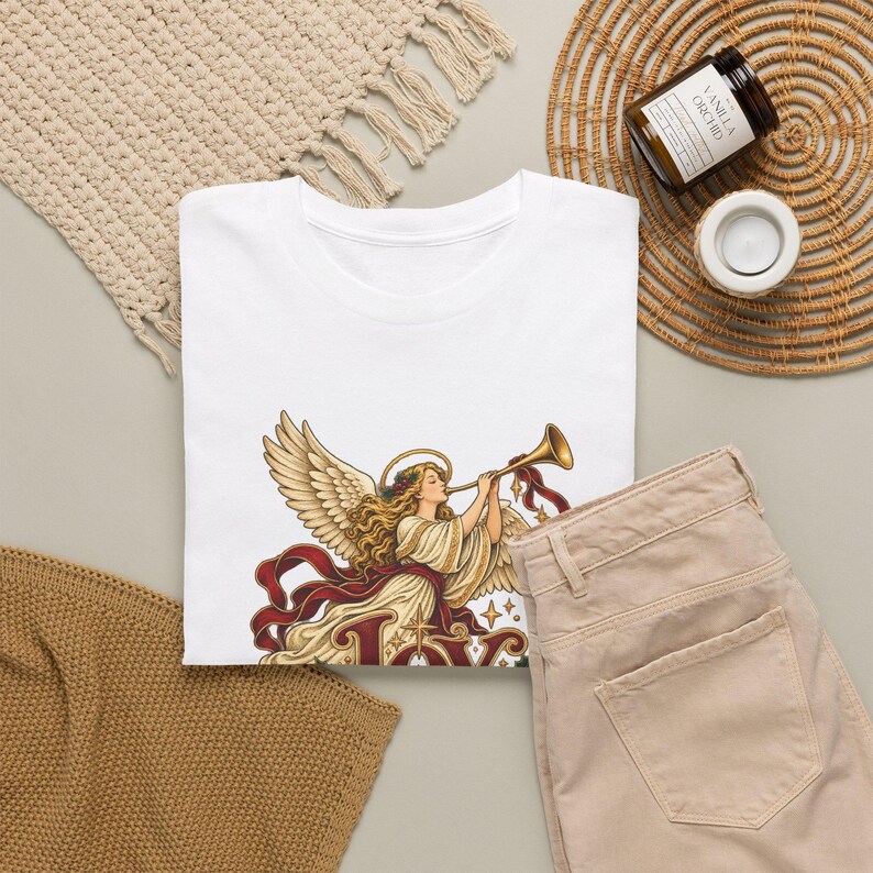 May include: A white t-shirt featuring a graphic illustration of an angel with large wings, long blonde hair, and a halo, playing a golden trumpet. Below the angel, the word JOY is written in large, decorative, dark red lettering.