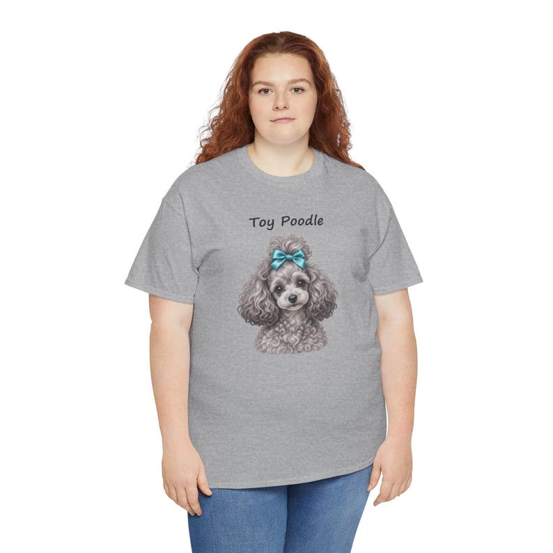 K&ouml;nnte beinhalten: Ein graumeliertes Kurzarm-T-Shirt mit einer Grafik eines lockigen, grauen Toypudels, der eine leuchtend blaue Schleife auf dem Kopf tr&auml;gt. Der Text "Toy Poodle" ist in schwarzen Buchstaben &uuml;ber der Hundeillustration auf der Vorderseite gedruckt.
