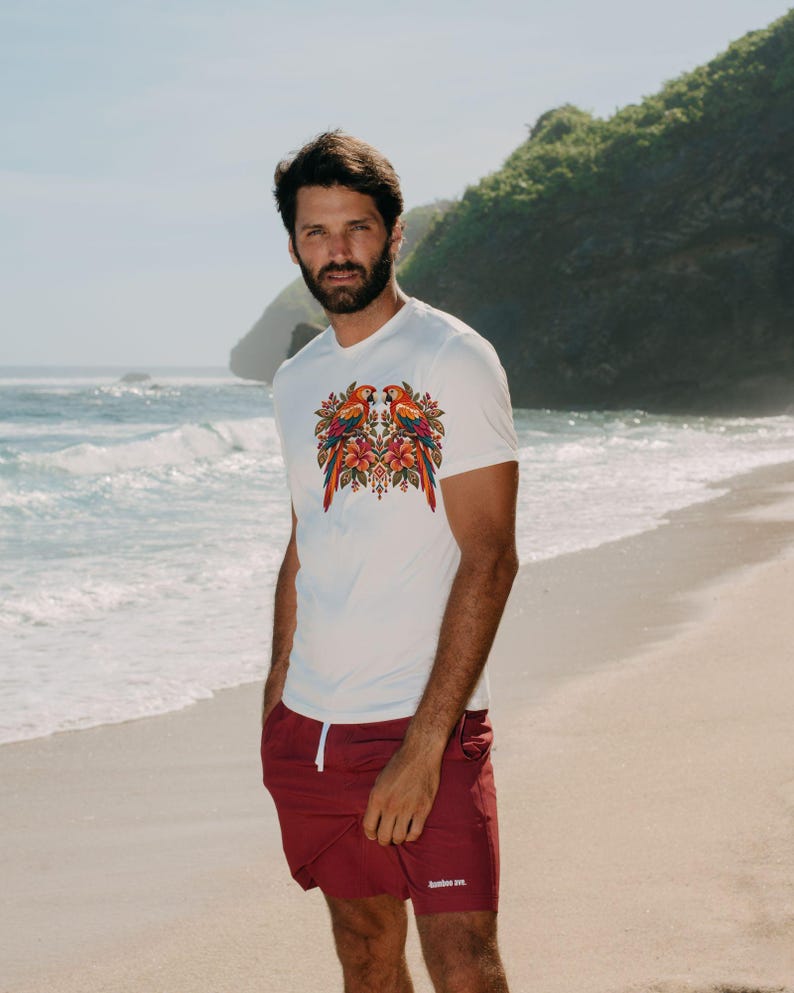 May include: A white short-sleeved t-shirt featuring a colorful graphic of two parrots surrounded by tropical flowers and foliage on the chest, paired with solid maroon shorts that have a white drawstring and small white text on the leg.