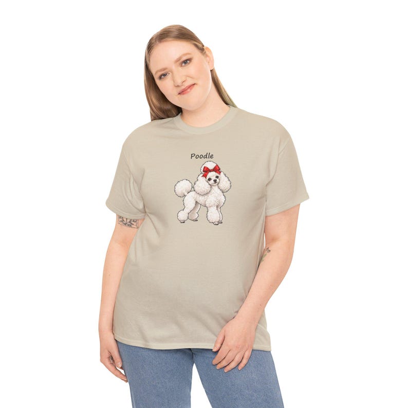 May include: A beige short-sleeved t-shirt featuring a graphic of a white poodle with a red bow on its head. The word Poodle is printed in black text above the dog illustration. The shirt has a crew neckline and a standard fit.