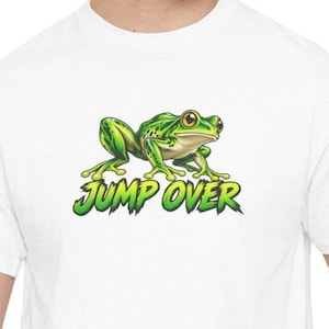May include: A white t-shirt featuring a graphic of a green frog with large eyes positioned above the text JUMP OVER in a stylized green font. The frog is depicted in a crouching stance with its legs spread out on the fabric.