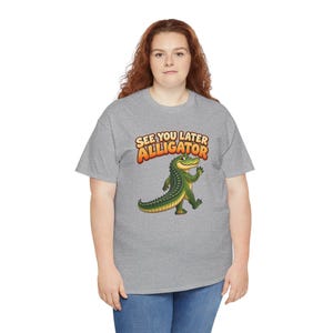 May include: A heather gray short-sleeve t-shirt featuring a cartoon green alligator waving its hand. Above the alligator, the text reads "SEE YOU LATER ALLIGATOR" in bold, orange, and yellow gradient lettering with a dark outline.