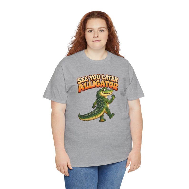 May include: A heather gray short-sleeve t-shirt featuring a cartoon green alligator waving its hand. Above the alligator, the text reads "SEE YOU LATER ALLIGATOR" in bold, orange, and yellow gradient lettering with a dark outline.
