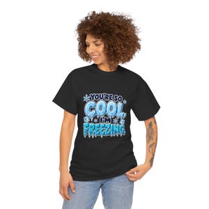 May include: A black short-sleeve t-shirt featuring a graphic print with the text "YOU'RE SO COOL, I'M FREEZING." The design includes light blue icy lettering, white snowflakes, and ice cubes, creating a winter-themed visual effect.