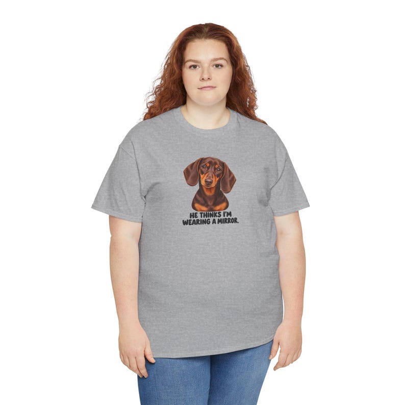 May include: A heather gray short-sleeve t-shirt featuring a graphic of a brown dachshund dog. Below the dog, the text reads: HE THINKS I'M WEARING A MIRROR. The shirt has a crew neckline and is worn with blue denim jeans.