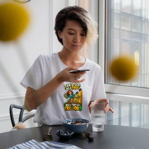 May include: A white short-sleeved t-shirt featuring a graphic print of a person with brown hair emerging from a tree trunk. The text reads I AM STUCK AS A TREE WHILE THEY GET TRILLIONS in green and yellow lettering with a small black bird.